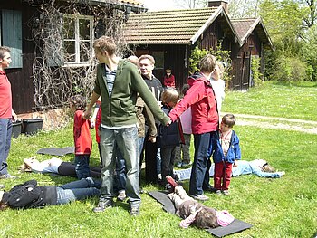 Eine Gruppe an Jugendlichen und Kindern stellt und legt sich Rücken an Rücken in eine Kreisformation, um einen Baum darzustellen