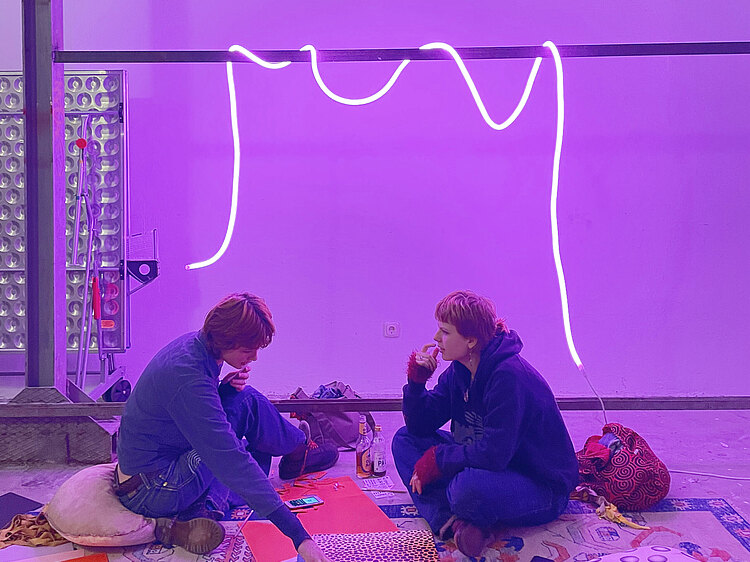 Zwei Menschen sitzen vor buntem Hintergrund am Boden und erschaffen Kunst