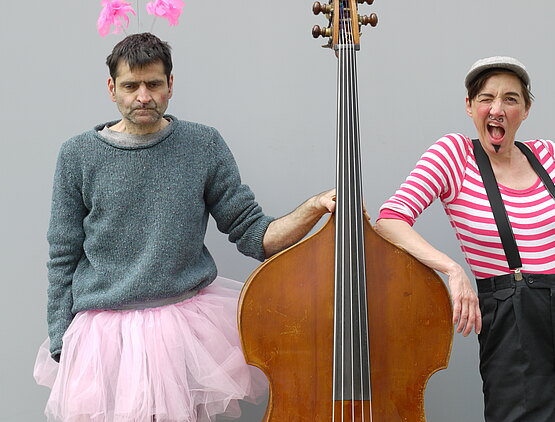 Zwei Schuaspieler die Klamotten getauscht haben posieren mit einem Cello