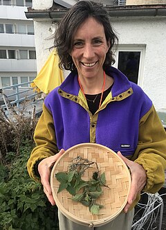 Auf dem Bild zu sehen ist Ulrike Zieher, die Ansprechperson der Community Dachgarten Werkstatt der Färberei. Sie steht auf dem Dachgarten und hält einen Korb voll geernteter Brennessel und anderer Kräuter in Händen.