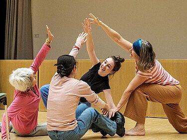Vier Frauen tanzen am Boden. Alle Körper haben unterschiedliche Formen, Ihre Arme bilden eine Art Pyramide. Man sieht deutlich dem Kontakt unter den Tanzenden.