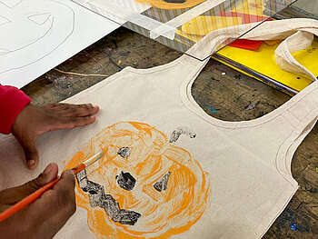 Kinderhände malen mit einem Pinsel einen Kürbis auf einen Stoffbeutel 