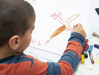Ein Kind malt mit Kreiden einen Vogel auf ein Blatt Papier. Vom Kind sieht man nur den Hinterkopf und die Schulter mit dem rechten Arm.