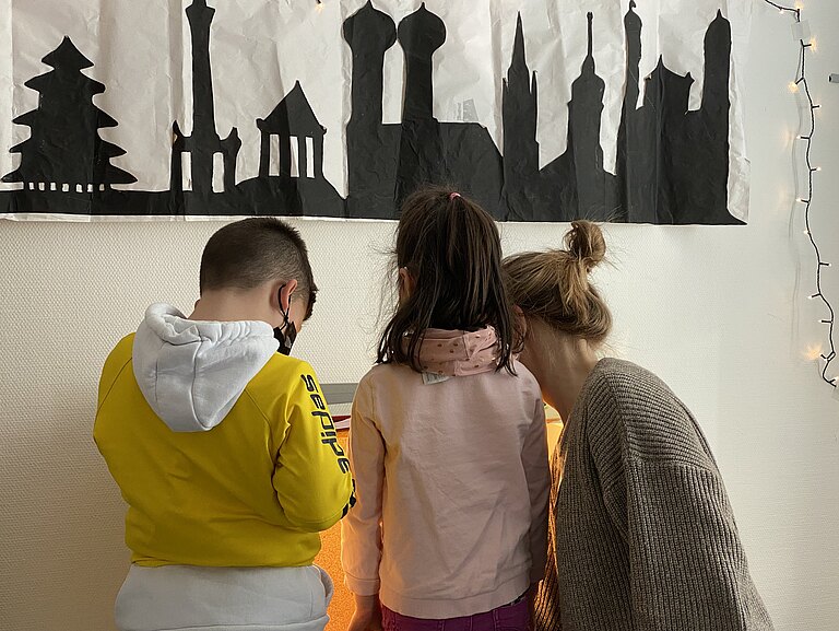 Drei Personen stehen vor einem an die Wand geklebten Scherenschnitt von Münchner Wahrzeichen. Sie wenden der Kamera den Rücken zu. Zwei davon sind Kinder.