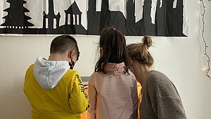 Drei Personen stehen vor einem an die Wand geklebten Scherenschnitt von Münchner Wahrzeichen. Sie wenden der Kamera den Rücken zu. Zwei davon sind Kinder.
