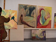 In einem weißen Atelierraum lehnen vier bunte Gemälde an der Wand. Im Vordergrund sitzt eine Person an einer Staffelei und malt mit einem Pinsel auf dem Bild, das auf der Staffelei steht.