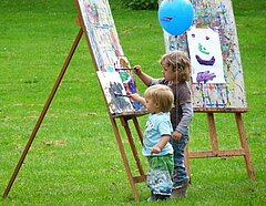 Zwei Kinder stehen auf der Wiese und malen gemeinsam ein Bild auf einer Staffelei