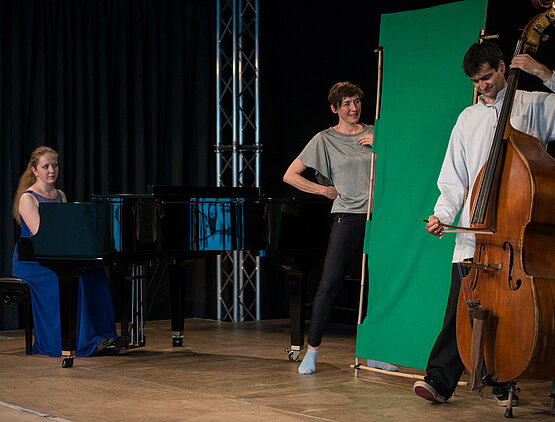 Ein Mann spielt Kontrabass, eine Frau spielt Klavier und eine Frau singt auf der Bühne