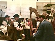Drei Musiker sitzen an einem Tisch mit älteren Gästen in einem Wirtshaus und spielen ihnen etwas vor