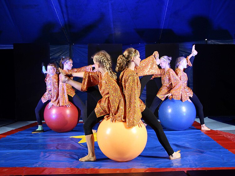 Sechs Mädchen in glitzernden  Kostüm zeigen eine Zirkusfigur auf Gymnastikbällen. Immer zwei Mädchen sitzen Rücken an Rücken auf einem Ball und strecken ein Bein hoch in die Luft.