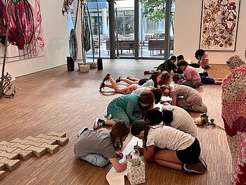 Kinder im Alter von ca. 11 Jahren beugen sich über ein am Boden liegendes, langes Papier und zeichnen konzentriert darauf. Sie befinden sich in mitten eines großen hellen Ausstellungsraums, in dem verschiedene angeschnittene Skulpturen zu sehen sind. Im Hintergrund hängt eine Blumenmalerei an der Wand.