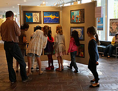 Mehrere Kinder unterschiedlichen Alters stehen vor einer Ausstellungswand, betrachten die gemalten Bilder und unterhalten sich über die Bildwerke.