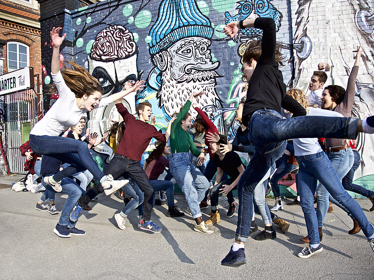 mehrer Jugendliche springen vor einer Wand mit Graffiti