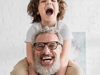 Ein älterer Herr hat seinen Enkel auf den Schultern sitzen, beide lachen