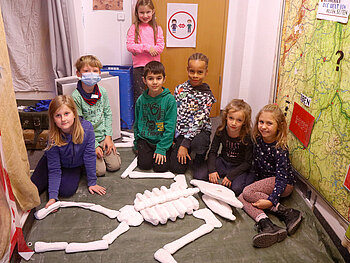Sechs Kinder sitzen auf einem Fußboden und vor ihnen liegt ein zusammengebautes Dinosaurier-Skelett