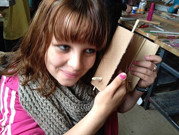 Eine Jugendliche hält sich ein aus einer Kartonschachtel selbst gebautes Instrument ans Ohr.