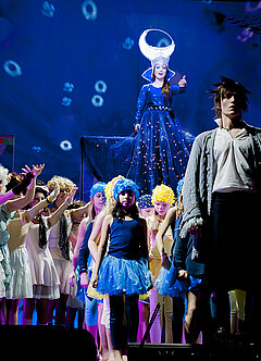 Im Hintergrund steht erhöht eine junge Frau in einem auffälligen blauen Kostüm und im Vordergrund ein junger Mann mit barock anmutender Jacke. Zwischen den beiden ist eine große Gruppe von Kindern und Jugendlichen in bunten Kostümen, die auf den Mann deuten oder blicken. Es handelt sich um eine Bühnensituation aus „Die Zauberflöte“.