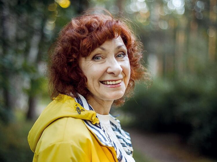 Ältere Dame mit roten lockigen Haaren steht im Wald und blickt lächelnd in die Kamera