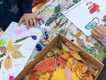 Auf einem buntem Tisch liegt einen Kiste mit Herbstblättern und Kinderhände bekleben weiße Papiere mit den Herbstblättern