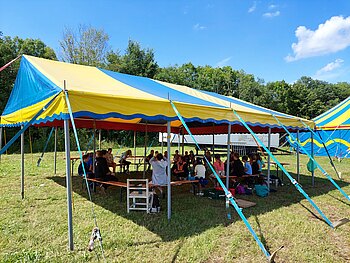Eine Gruppe Personen sitzt in einem blau-gelb gestreiften Zelt im Kreis. Im Hintergrund ist ein blau-gelb gestreiftes Zirkuszelt.