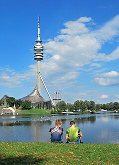 2 Buben sitzen am See im Olympiapark und schauen auf den Turm am gegenüberliegenden Ufer
