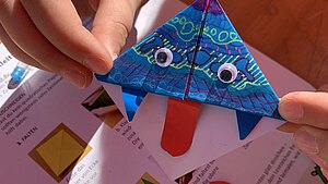 Ein Lesezeichen, das aus Origamipapier gefaltet wird. Es ist mit Mustern, angeklebten Augen und einer Zunge verziert.
