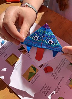 Ein Lesezeichen, das aus Origamipapier gefaltet wird. Es ist mit Mustern, angeklebten Augen und einer Zunge verziert.
