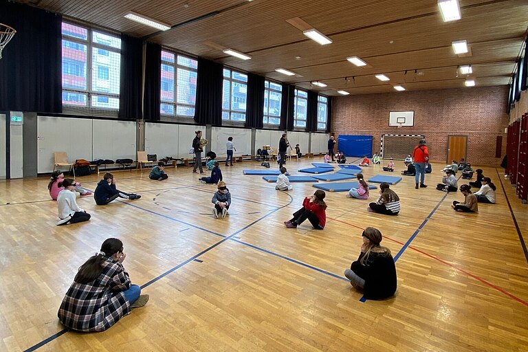 Die Kinder einer Schulklasse sitzen mit verbundene Augen auf dem Boden einer Turnhalle und hören Instrumenten zu.