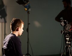 Foto zeigt einen Jugendlichen der auf einem Stuhl sitzt und in eine Filmkamera schaut