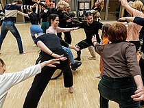 Viele tanzende Menschen unterschiedlichsten Alters und unterschiedlicher Körperlichkeiten bilden einen Pulk, bei dem langgestreckte Arme und Beine die Tanzenden verbinden.