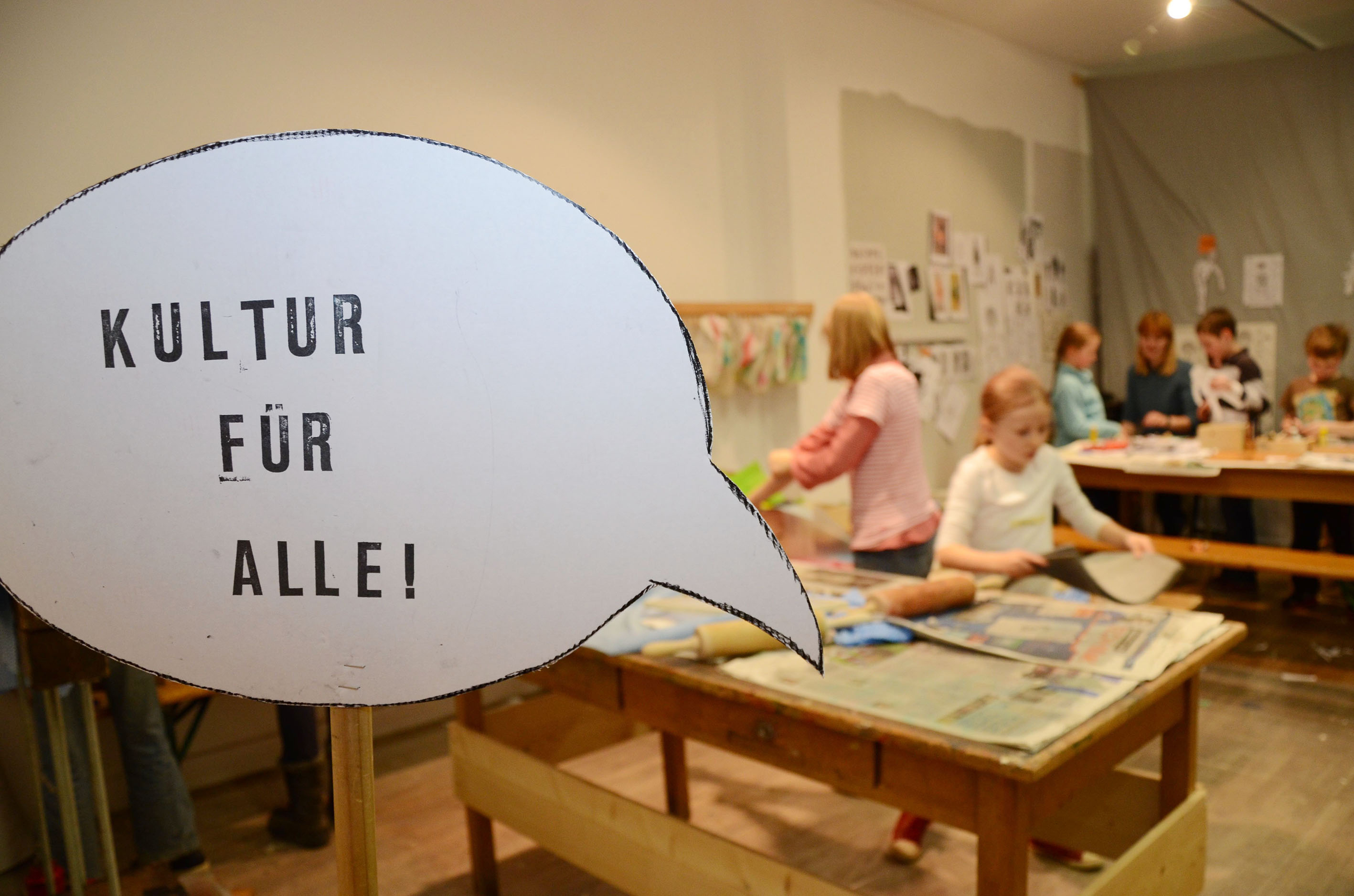 Der Text "Kultur für alle!" steht in einer Sprechblase, im Hintergrund sieht man Kinder in einer Buchwerkstatt. 